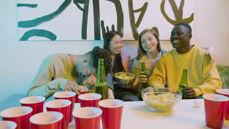 happy multiethnic young friends drinking beer, eating potato chips and talking while sitting on sofa at home.