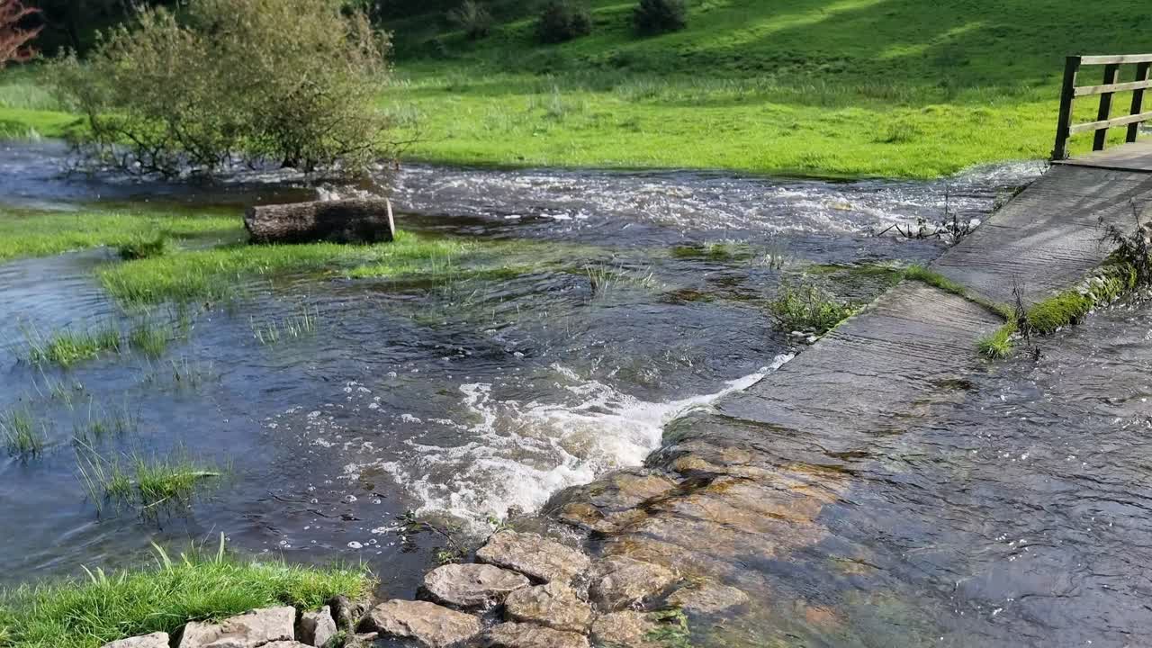 desbordando la orilla del río inundando el pacífico norte de gales iluminado por el sol prado bajo un puente de madera cruzando el camino