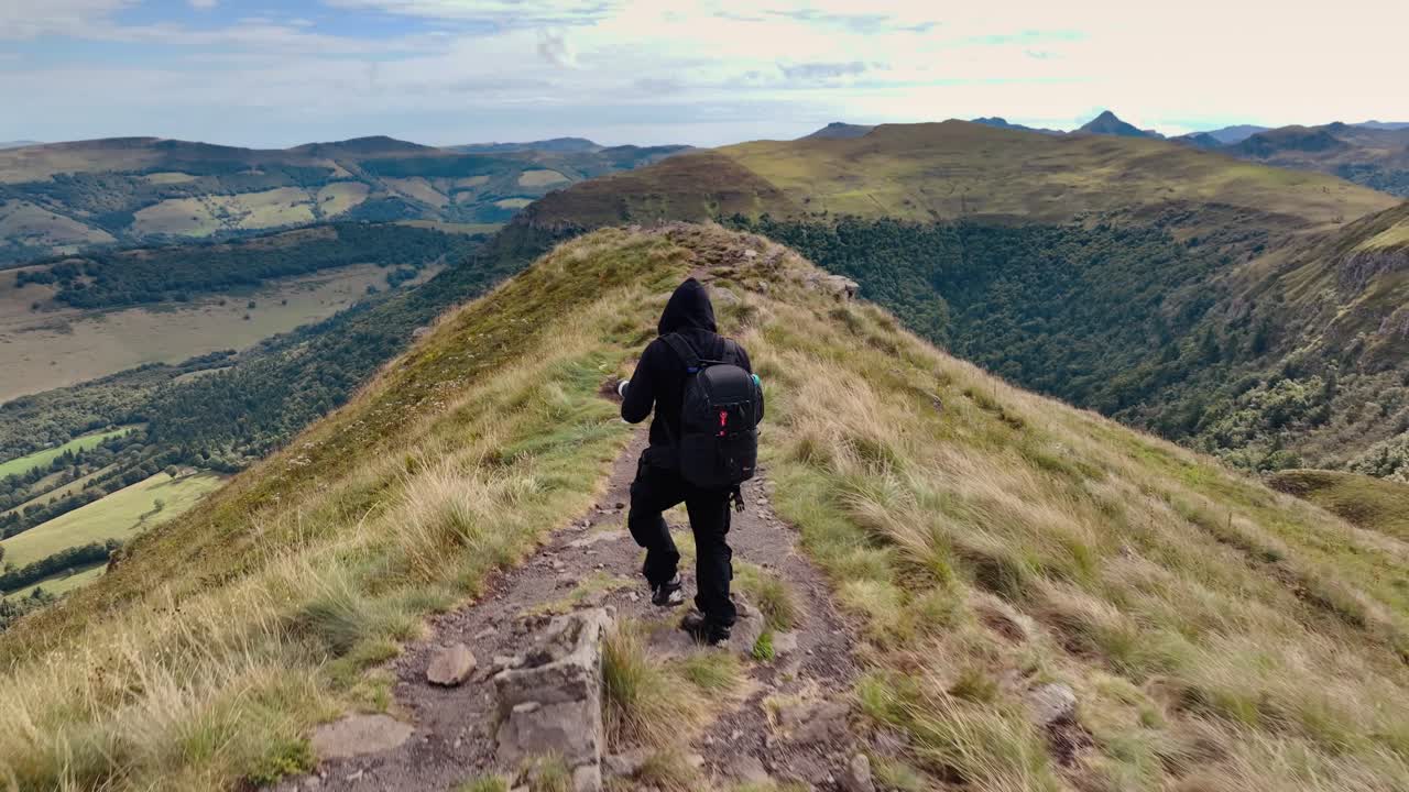 Flygfoto av en fotograf med ryggsäck som står på en smal bergskam i Auvergne-Rhône-Alpes och fotograferar dalen