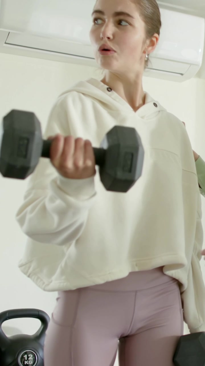 Vertical video: Following cue for form, hooded woman curling hex dumbbell home, coach guiding wrist