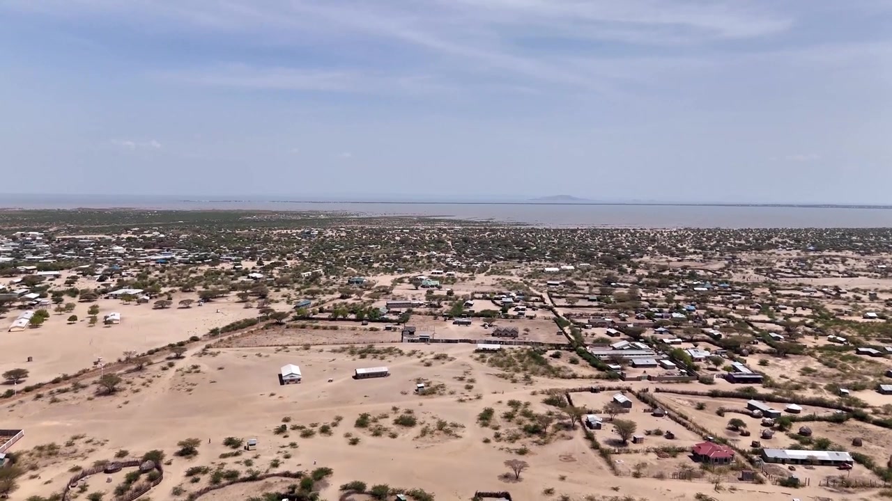 Laaja dronekuva ilmasta laajasta, pölyisestä asutuksesta lähellä Turkana-järven rantoja Keniassa, jossa on hajallaan rakennuksia, aitoja ja kuivaa maastoa järven siintäessä horisontissa