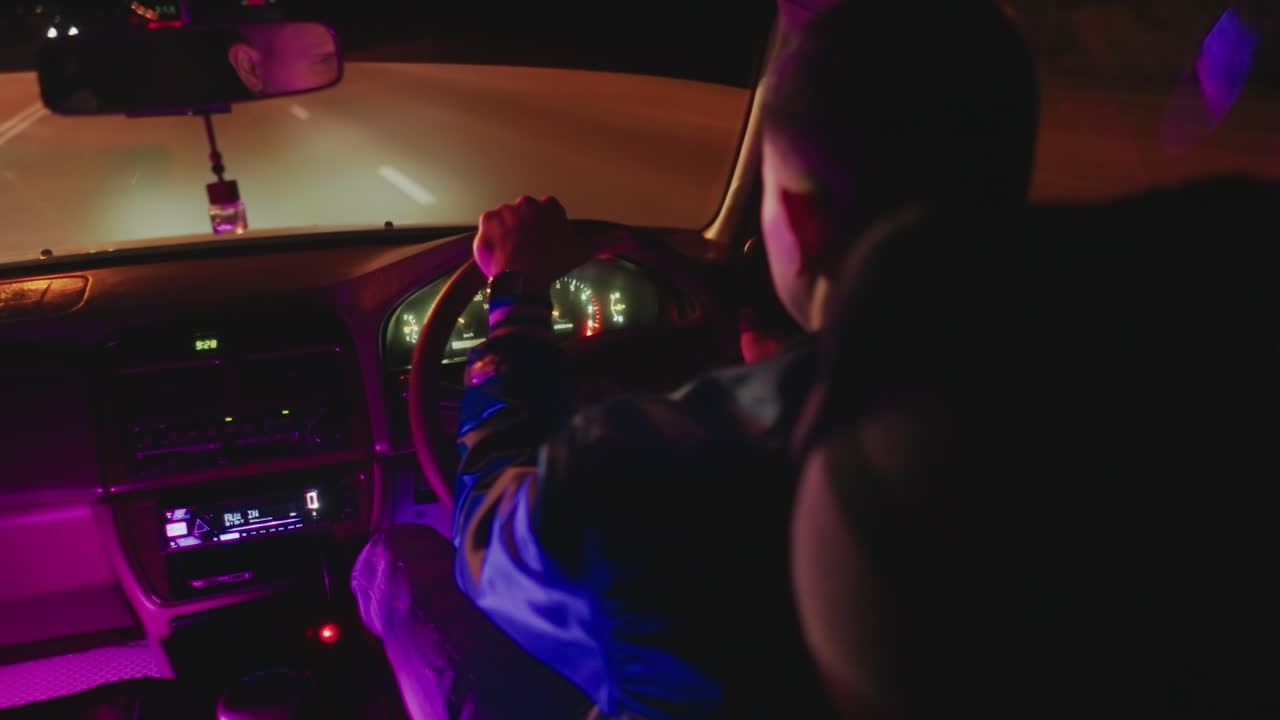 vista interior nocturna del conductor con las manos en el volante, brillo neón púrpura y azul en el salpicadero, velocímetro iluminado, parabrisas enmarcando la carretera vacía, espejo retrovisor reflejando luces distantes
