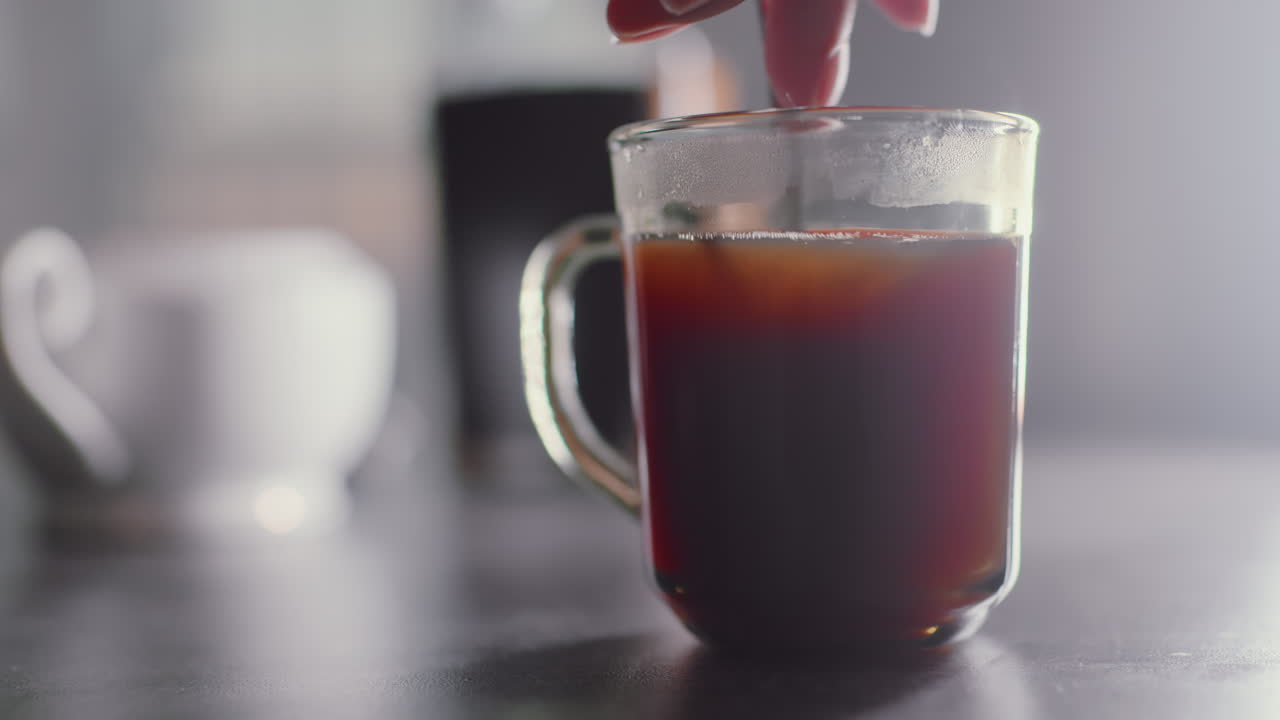 Close up of individual hand stirring coffee inside clear glass mug, steam rising from hot dark liquid while spoon swirls, morning beverage preparation scene with blurred background cup visible