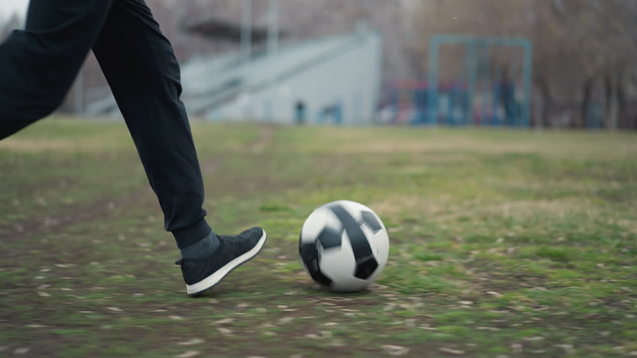 Close-up of someone in black pants and sneakers running with a soccer ball on a grassy field, with a blurred background showing people moving around and buildings
