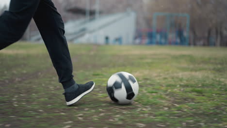 primer plano de alguien con pantalones negros y zapatillas de deporte corriendo con una pelota de fútbol en un campo cubierto de hierba, con un fondo borroso que muestra a la gente moviéndose y edificios