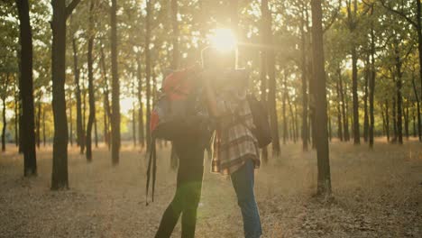 A-happy-blonde-girl-with-a-bob-hairstyle-in-a-plaid-shirt-with-a-large-red-hiking-backpack-and-a-brunette-girl-with-a-red-bandana-meet-during-a-hike-and-hug-against-the-backdrop-of-a-summer-sunny-forest