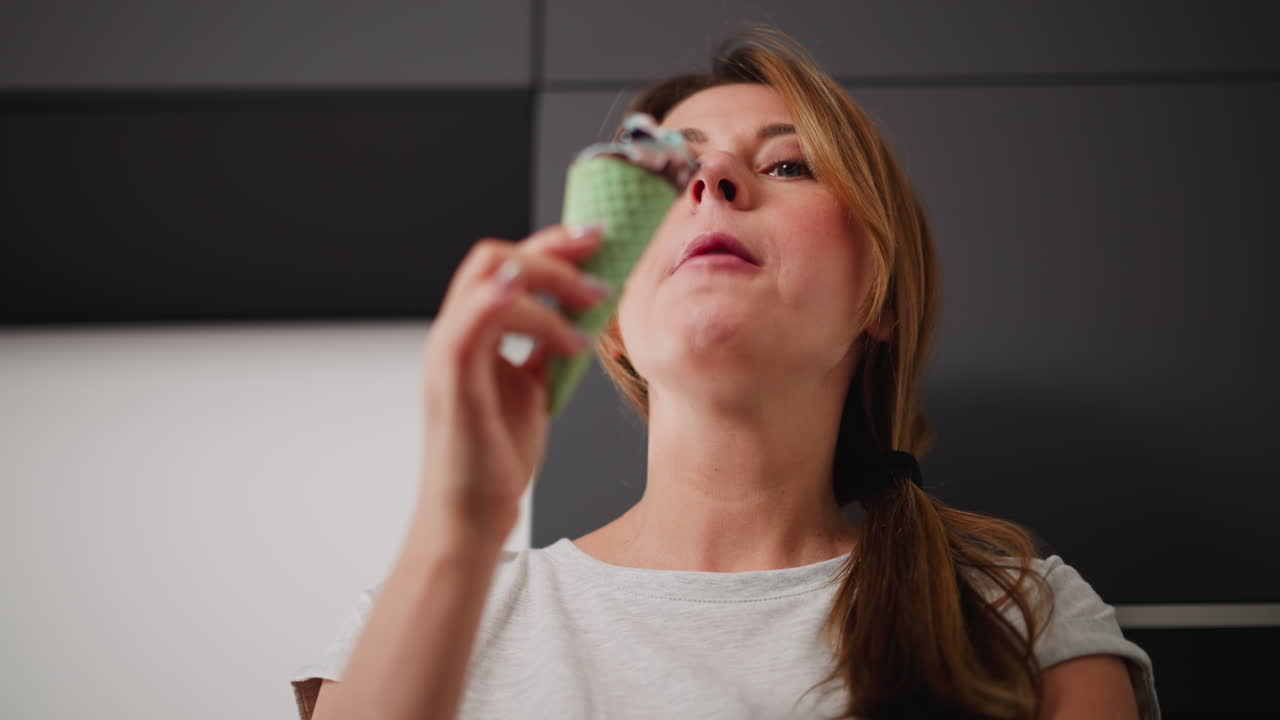 Close up of housewife with folded arm savoring colorful ice cream cone with green waffle cone, glancing thoughtfully at sweet treat, dressed casually in light grey t-shirt