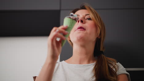 close up of housewife with folded arm savoring colorful ice cream cone with green waffle cone, glancing thoughtfully at sweet treat, dressed casually in light grey t-shirt