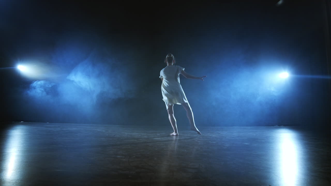 Dramatic spins and flips of a modern ballet dancer from a musical. A single woman emotionally dances on stage against a dark background with smoke in the spotlight in slow motion.
