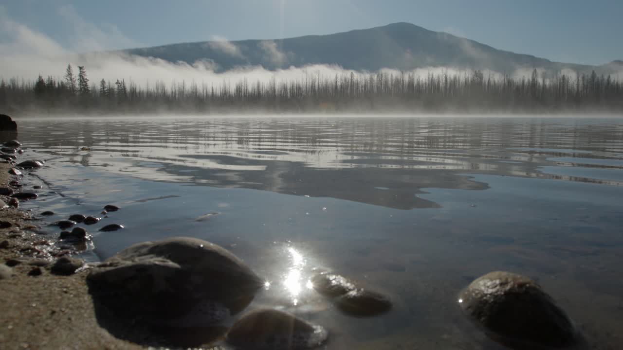 Calm mountain lake at sunrise with mist rising from the water. Trees line the shoreline and a distant mountain is reflected on the lake beneath a clear morning sky