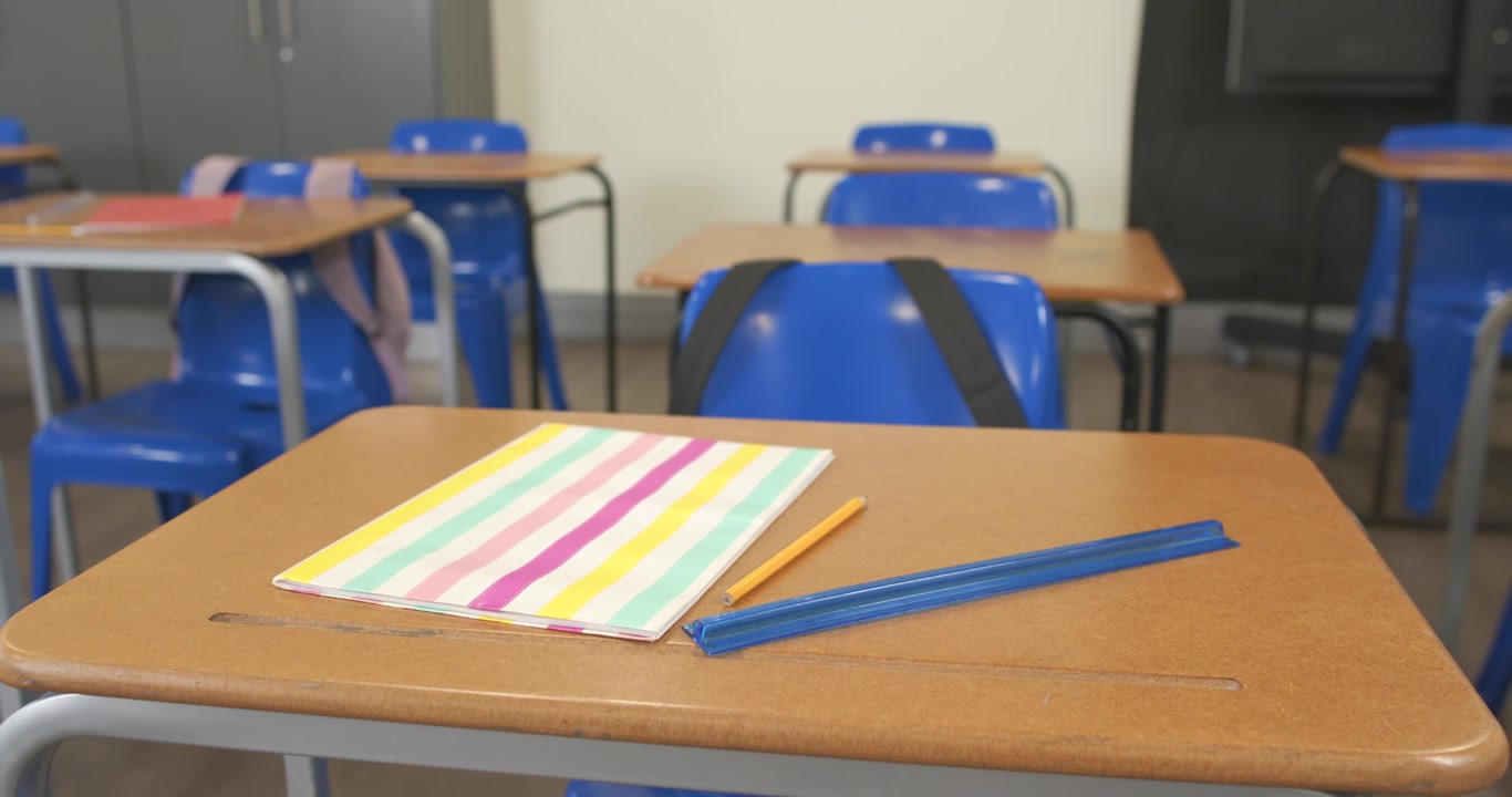La cámara se desplaza y reenfoca cerca de un pupitre de aula para resaltar un cuaderno pastel, un lápiz y un estuche.