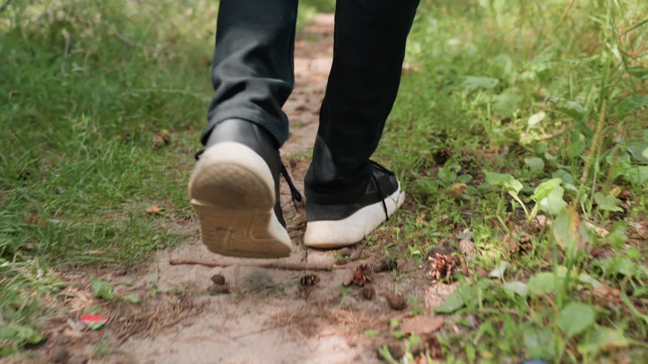 Back leg view of man in black trousers and canvas sneakers walking calmly on forest pathway surrounded by green plants, grass, and soil in peaceful natural outdoor environment during daylight