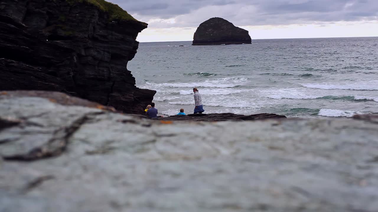 Family of four is sitting on a rocky coast next to a mountain in the evening. There is a woman, man and children. Tide is high and beach is covered in water. Sun has set, clouds are grey. Filmed in HD