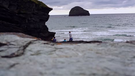 family of four is sitting on a rocky coast next to a mountain in the evening. there is a woman, man and children. tide is high and beach is covered in water. sun has set, clouds are grey. filmed in hd