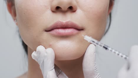 hands in gloves giving injection in lip of asian woman on grey background, slow motion