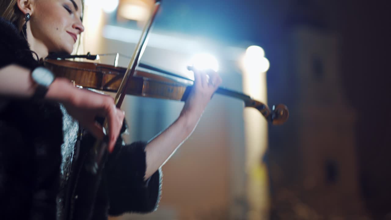 A creative girl in a black coat is playing the violin a romantic melody on the background of light from an evening lantern in the city. Close-up