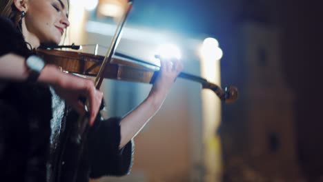 a creative girl in a black coat is playing the violin a romantic melody on the background of light from an evening lantern in the city. close-up
