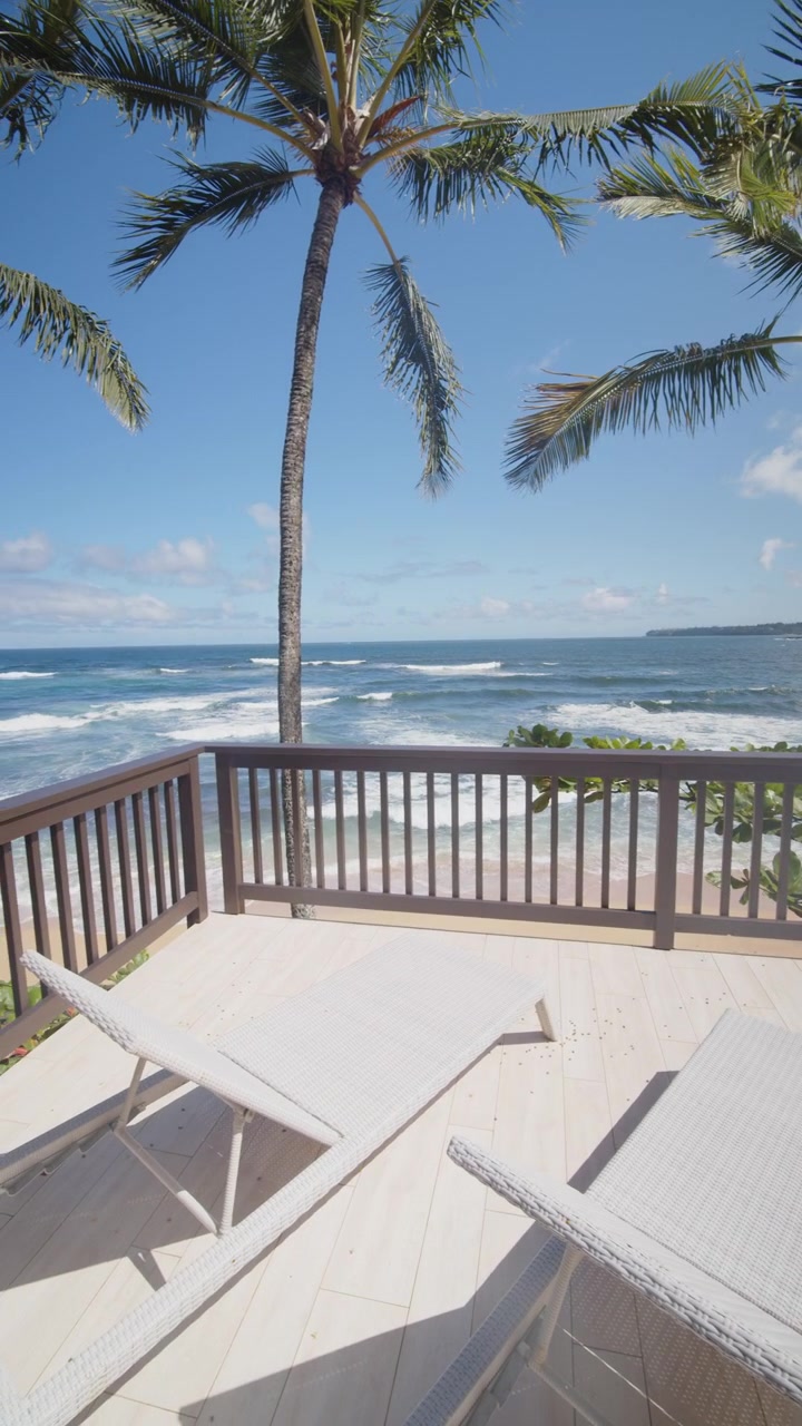 Palm trees sway above an oceanfront deck with lounge chairs in Kauai, Hawaii.