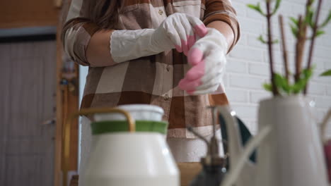 lady tucking single white rubber glove with pink fingertips on hand while standing by indoor winter plants and brick wall, preparing gentle plant care in cozy home greenhouse environment