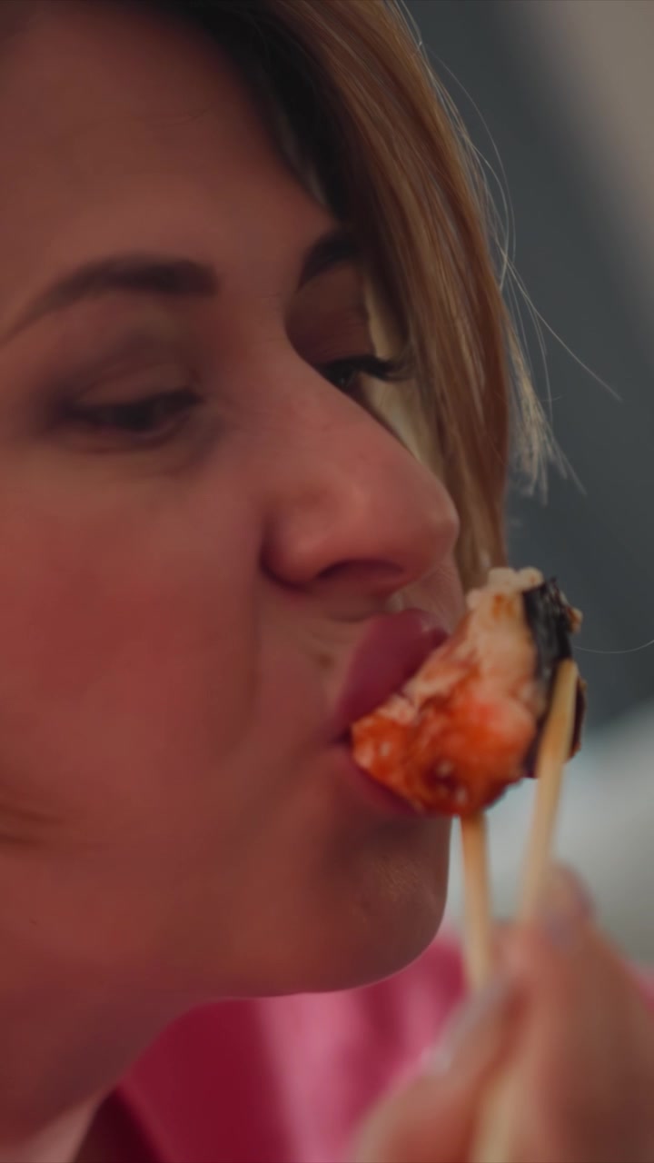 Close up of woman eating sushi with chopsticks bringing food to mouth and chewing. Background softly blurred emphasizing action in soft lighting closeup