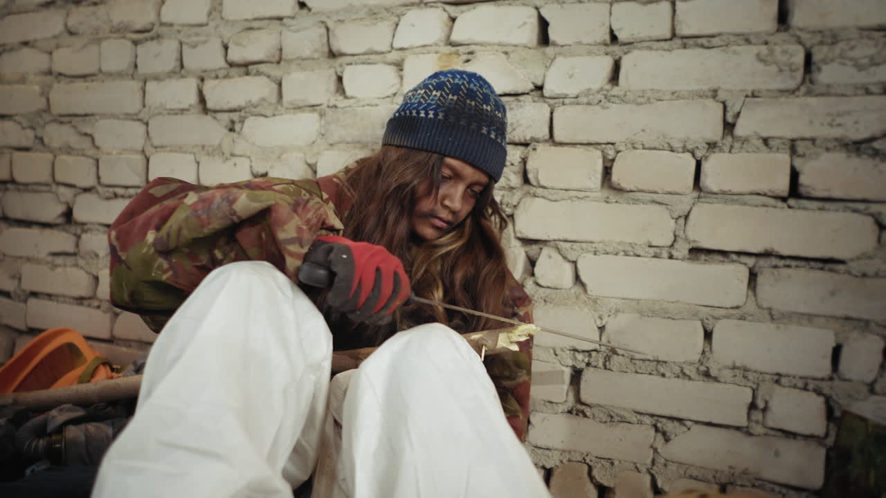 겨울 모자와 더러운 장갑을 낀 버려진 아이가 차가운 폐허 건물 안 벽돌 벽에 기대어 앉아 컷러스로 무기를 조각하며, 고난 속에서 생존을 위해 집중하고 있어요.