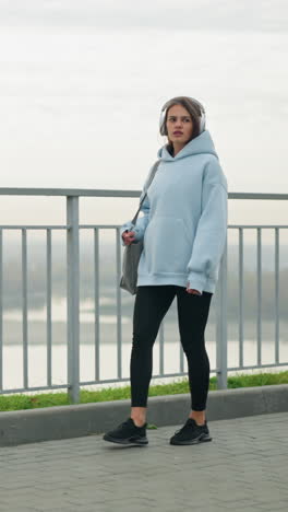 fair woman in casual wear walking slowly while listening to music with headphones, holding a bag, with water and green grass in the background, creating a serene and relaxed atmosphere