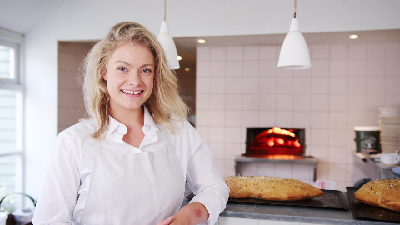 Young female business owner stands leaning at the counter of an artisan bakery looking to camera, beside freshly baked loaves of bread, close up