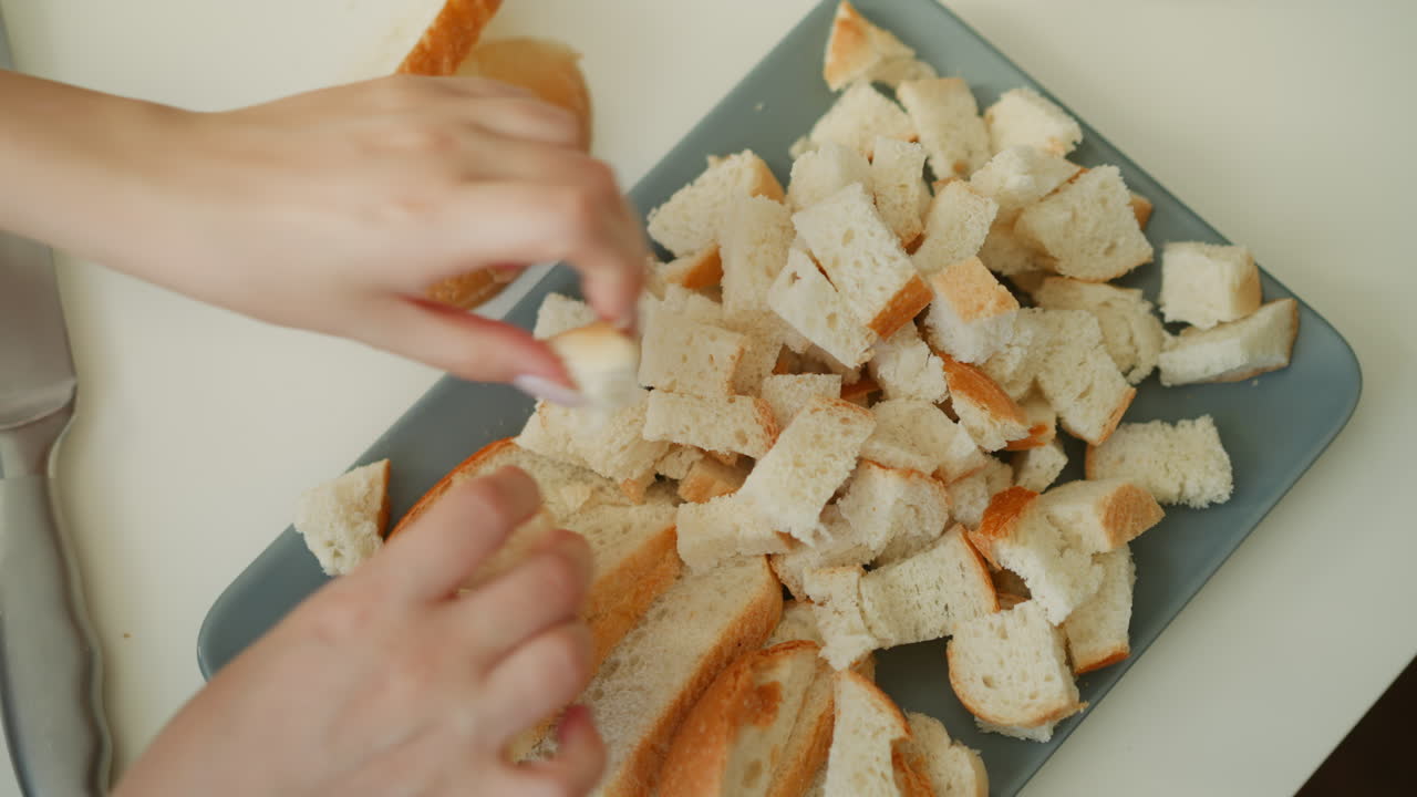 Pastry chef hand placing sliced bread and small cubes on plastic tray under soft window light, crumbs and loaf ends on white table, capturing precise calm culinary preparation moment
