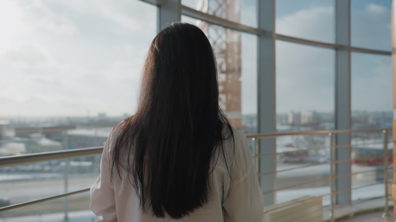 Back view of woman with straight long hair wearing white shirt standing by glass railing and gazing outdoors at busy urban scenery under cloudy sky with city buildings and traffic in soft daylight