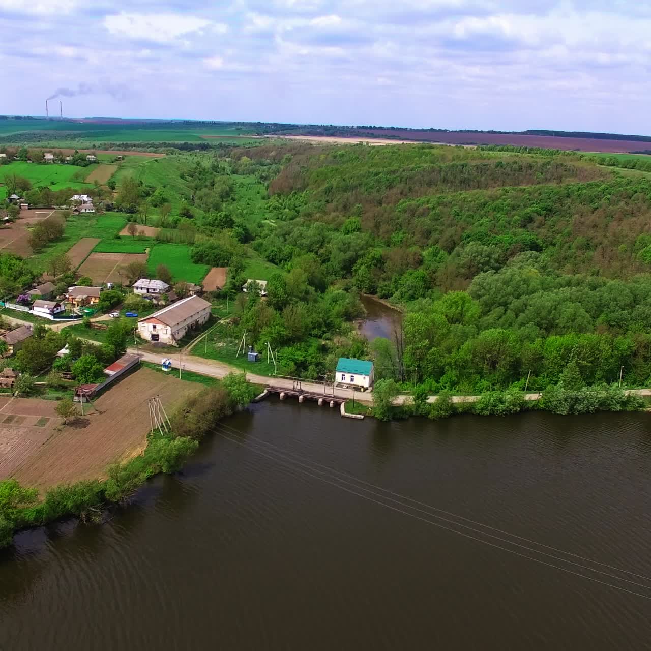 Dark waters of the river neighboring the village. Beautiful countryside along with green forests and farmlands at backdrop. View from above