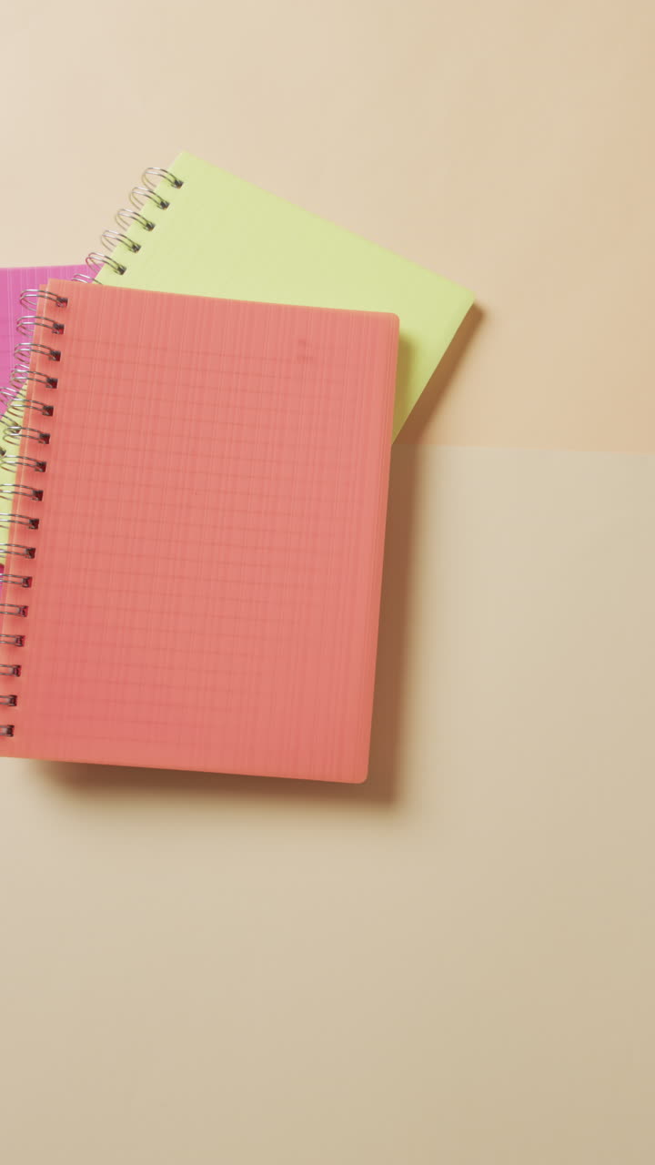 Vertical video of overhead view of colourful notebooks on beige background, in slow motion