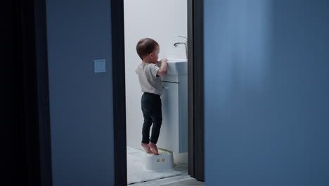 baby boy stands on toes to open water tap. kid is happy to watch the water flow.