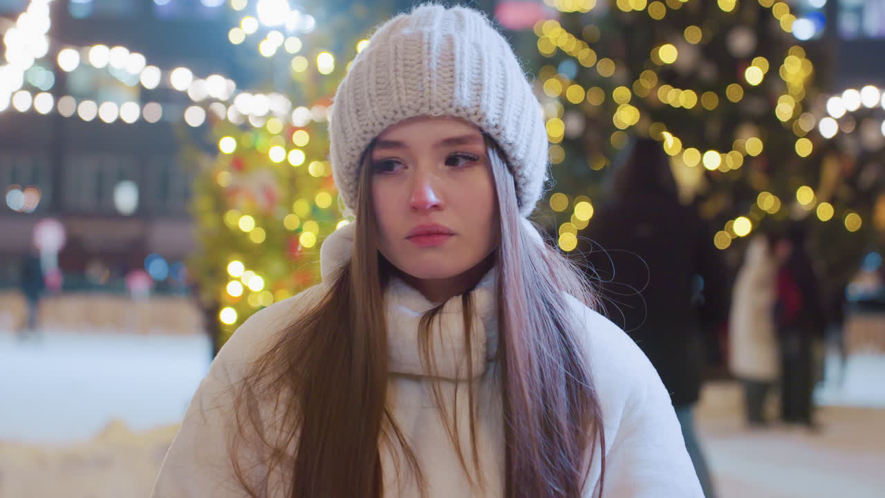 Young woman in cozy winter outfit sits outdoors at night, looking pensive, festive city lights illuminate the background as people skate, socialize, and enjoy the lively winter atmosphere around her