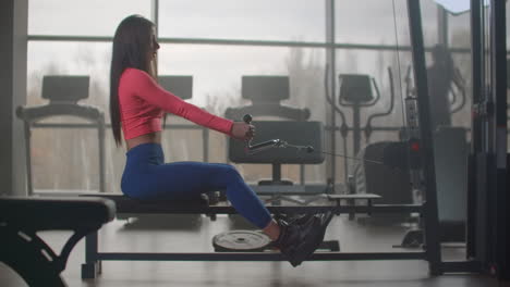 the young woman pumps her back and shoulder blades on the training machine. she pulls the handle to the lower abdomen on exhalation and returns to its original position on inhalation.