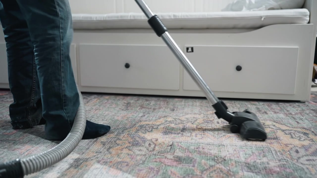 Široký záběr vysavače na vzorovaném koberci. Klidná domácí scéna pomalých tahů vysavače a stabilního rytmu, terapeutická nálada při úklidu s měkkým světlem, uklizený obývací prostor, viditelná hadice a modrá nádoba.