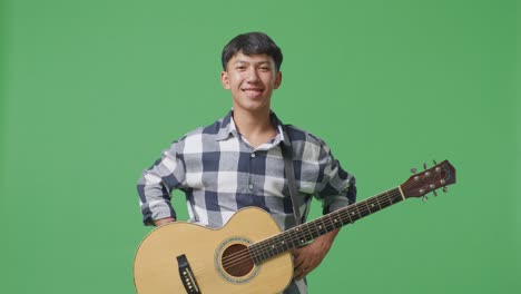 기타를 들고 있는 아시아인 십대가 이에 손을 고 녹색 화면 배경에 미소 짓고 있다.