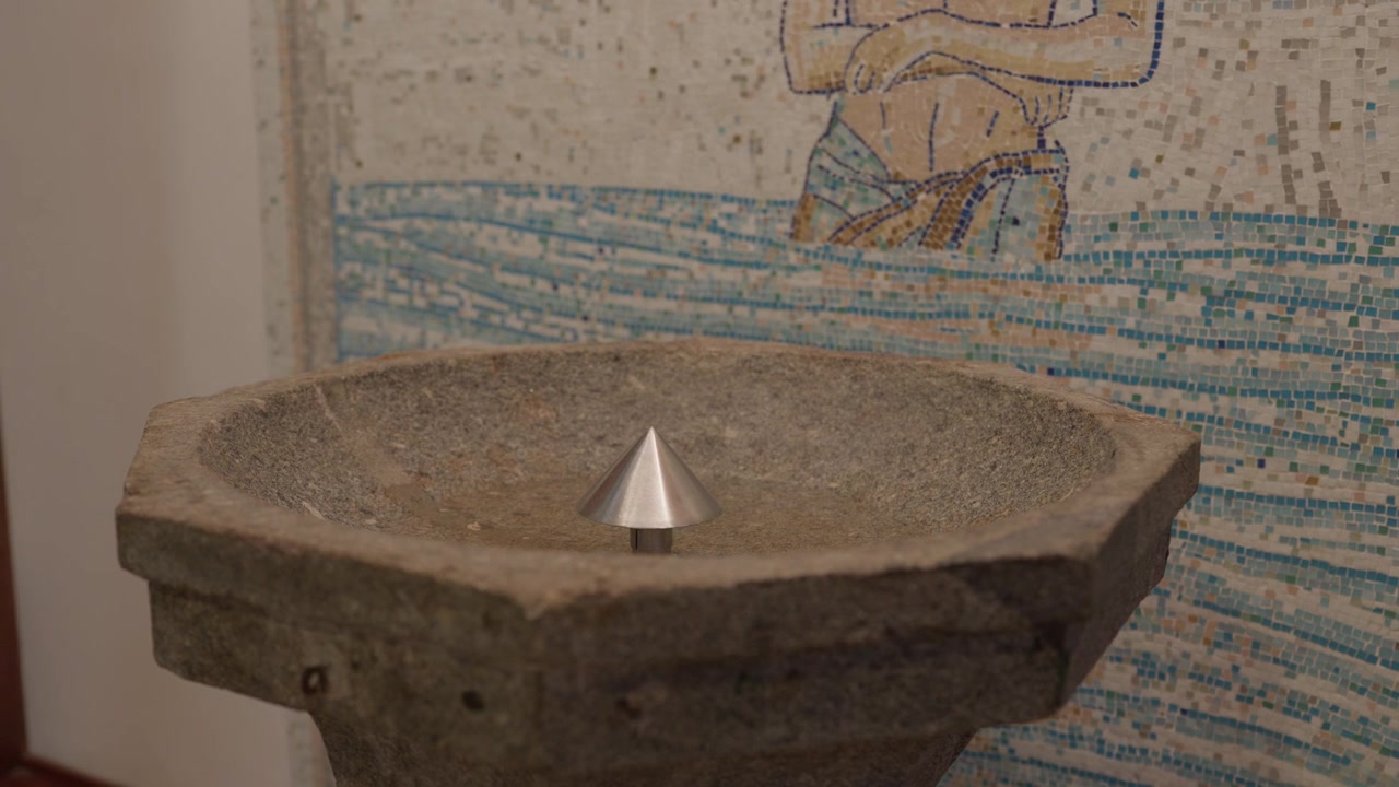 Fonte battesimale ottagonale in pietra vuota con un cono di metallo contro una parete a mosaico blu.