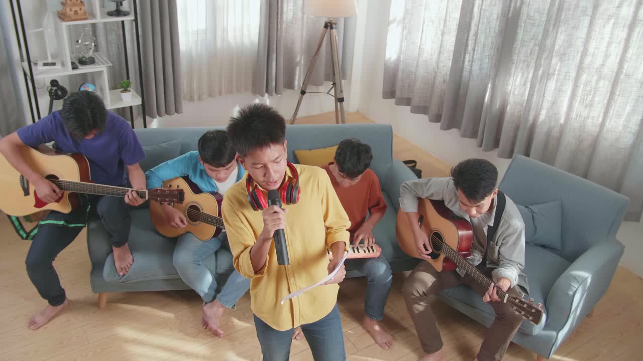 어린 아시아 소년이 노래를 부르고 종이에서 가사를 읽으며 그의 친구들은 집에서 소파에서 악기를 연주합니다.
