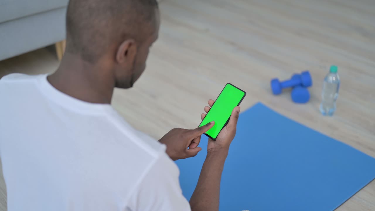 vista trasera de un hombre africano usando un teléfono inteligente con pantalla de tecla de croma en una alfombra de yoga