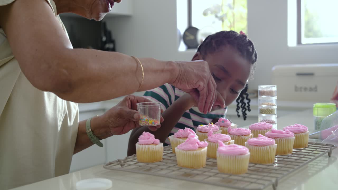 Donna anziana afroamericana che insegna a una ragazza a decorare cupcake con codette di zucchero in cucina.