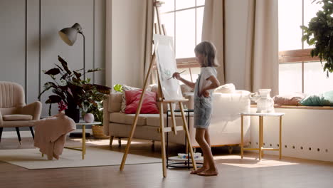 Rear-View-Of-A-Blonde-Girl-Painting-A-Plant-With-A-Brush-On-A-Lectern-In-The-Living-Room-At-Home