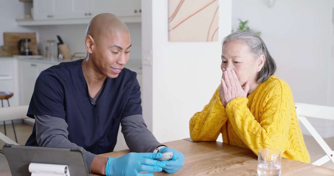 Badante afroamericana che controlla la glicemia di una donna anziana asiatica durante un controllo in cucina con un glucometro.