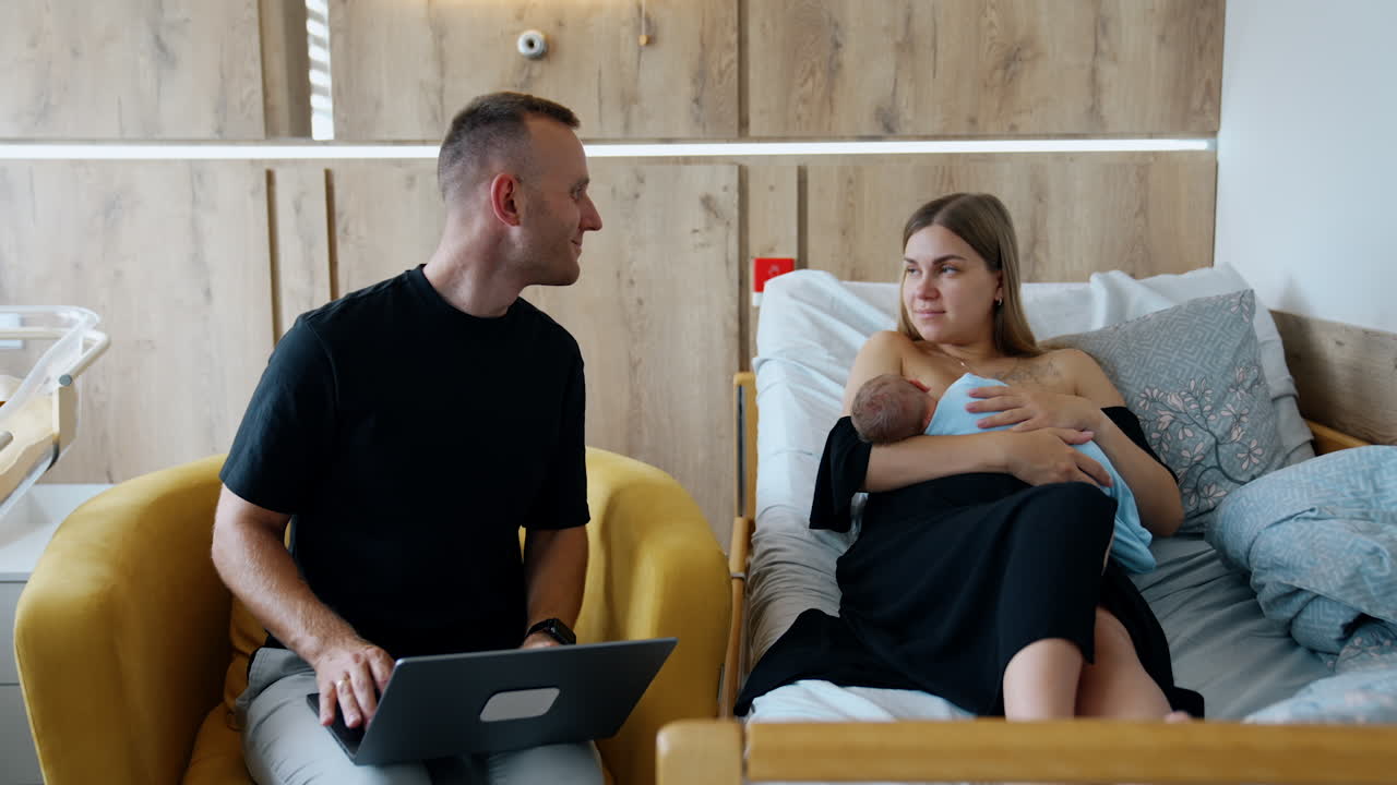 New family of three in the maternity hospital. Mother breastfeeding a newborn and man sits beside holding a laptop.