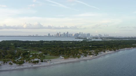 Trucking-right-aerial-drone-shot-of-the-beautiful-tropical-beach-surrounded-by-palm-trees-on-Crandon-Park-in-Key-Biscayne-with-the-skyline-of-Miami,-Florida-in-the-distance-on-a-sunny-summer-evening