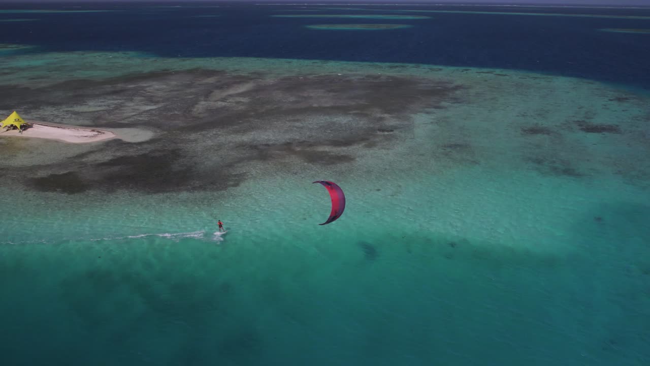 surfista de cometas rojo deslizándose sobre aguas turquesas cerca de una isla de arena, vista aérea