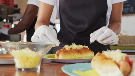 Mid-section-of-african-american-woman-wearing-apron-putting-grated-cheese-over-hot-dog-in-food-truck