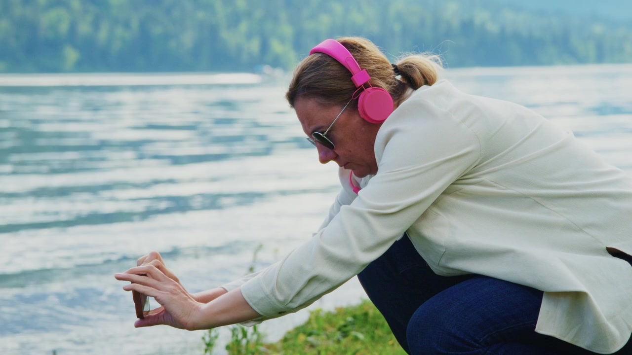 Una donna si accovaccia vicino al lago, tenendo il telefono per immortalare la natura. Indossa cuffie rosa, occhiali da sole, concentrata sull'acqua. Rilassata, pacifica, si gode il momento all'aperto. Perfetto per viaggi, benessere.