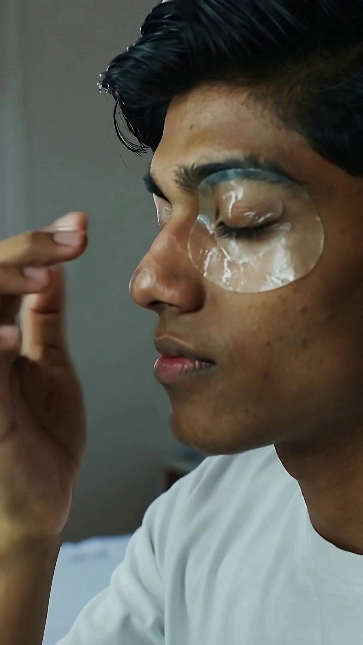 Vertical video: Holding phone man in tee on bed applying, pressing gel patch under eye for skincare