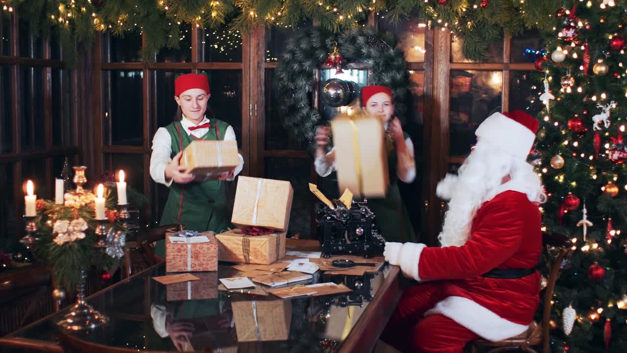 Christmas elves stack New Year's gifts on a Santa Claus table in a room with a Christmas tree and decorations
