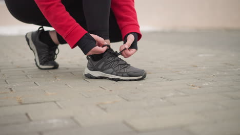 side view of athlete tying her right shoelace while squatting on interlocked path with blurred snow-covered background, depicts focused preparation for outdoor activity in winter